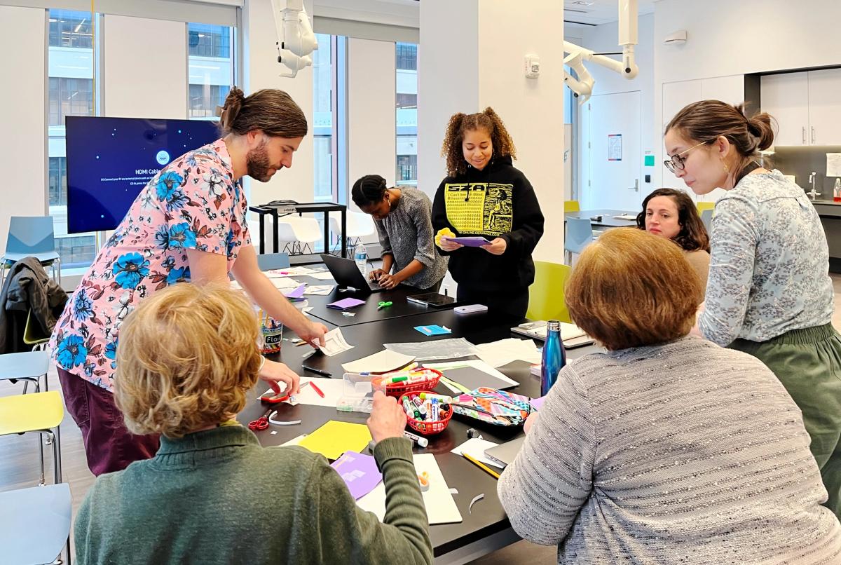 A group of eight people gathered around a rectangular table in a bright, modern classroom. They are engaged in a hands-on activity involving colorful paper, markers, scissors, and other craft supplies. One person is standing and helping others, while others are seated or reading instructions. A large screen in the background displays a message about an HDMI cable, and large windows bring in natural light from a cityscape outside.